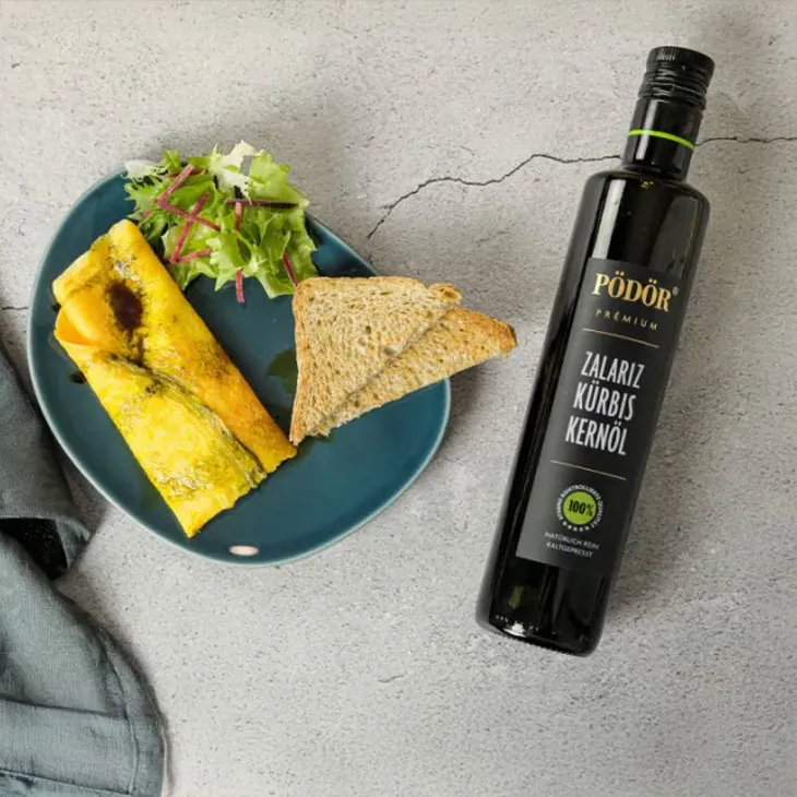 Flasche Pödör Zalariz Kürbiskernöl neben einem Teller mit Omelett, Salat und Vollkornbrot.